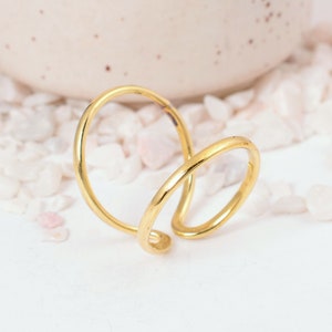 Puede incluir: Un anillo dorado con un diseño abierto único. El anillo presenta una forma curva y fluida que se envuelve alrededor del dedo. El anillo está sobre un fondo blanco con pequeñas piedras de color claro.