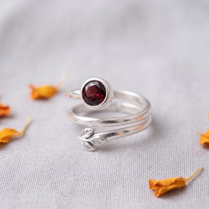 May include: A silver bypass ring featuring a round, faceted dark red garnet gemstone set in a bezel at one end and a small, sculpted leaf motif at the other end. The band has a smooth, polished finish and an open, adjustable design.