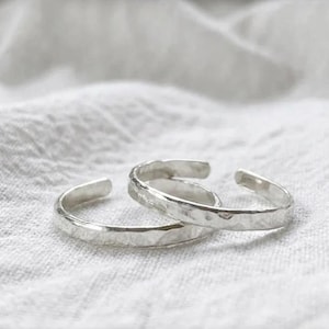 Könnte beinhalten: Zwei silberfarbene offene Ringe mit einer gehämmerten, strukturierten Oberfläche. Die Ringe sind kreisförmig mit einer kleinen Lücke und weisen ein metallisches Finish sowie ein reflektierendes, unebenes Außendesign auf.