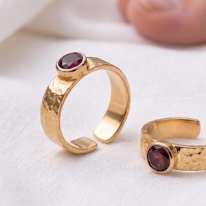 Peut inclure: Deux bagues ouvertes dorées présentant une texture martelée sur l'anneau et une pierre précieuse grenat ronde d'un rouge profond sertie dans une monture. Les bagues sont ajustables avec une ouverture à l'arrière, offrant une finition polie.