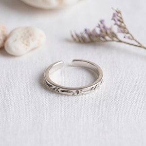 Puede incluir: Un anillo abierto de color plateado con un acabado pulido y una serie de pequeños patrones grabados en forma de hojas o enredaderas en la superficie exterior. El anillo tiene una abertura en la parte superior, lo que permite ajustar su tamaño.