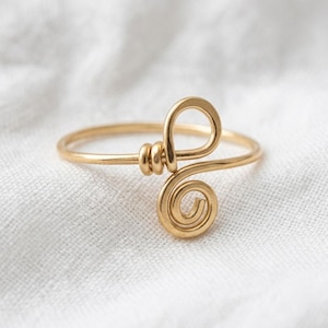May include: A gold-toned wire ring featuring a central design composed of a small circular coil and a larger looped wire element. The band is thin and smooth, with the decorative wirework securely wrapped around the main circular ring structure.