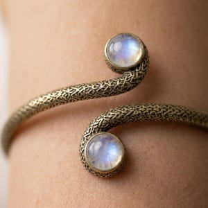 May include: An open cuff bracelet featuring a textured, dark metallic band with an intricate, woven-like pattern. The two ends of the bracelet terminate in circular settings, each holding a smooth, round, iridescent moonstone cabochon.