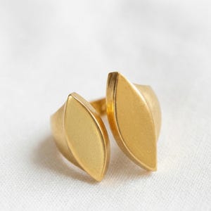 Op de afbeelding: Een goudkleurige ring met een bladachtig ontwerp. De ring heeft twee gebogen, langwerpige vormen die in het midden samenkomen en een open ontwerp creëren. De ring staat tegen een witte achtergrond.