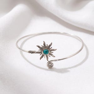 May include: A sterling silver cuff bracelet featuring a sun-shaped centerpiece with a round turquoise stone at its center. The metal band has a polished finish, a spiral end, and is clearly stamped with the number 925 to indicate silver purity.