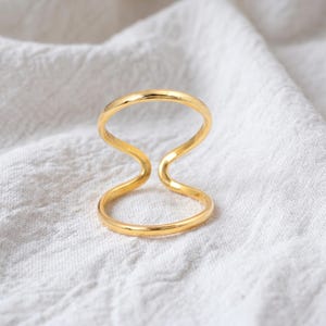 Könnte beinhalten: Ein goldfarbener Ring mit einer glatten, durchgehenden Ringschiene, die sich zu einer offenen Acht formt. Das Metall hat eine polierte, reflektierende Oberfläche und ein schlichtes Design, das zwei miteinander verbundene Schlaufen bildet.