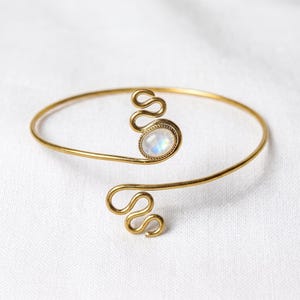May include: A gold-toned open cuff bracelet featuring a smooth, oval-shaped iridescent moonstone cabochon set in a decorative frame. The bracelet ends are finished with a distinct, swirling wire design that curves into a serpentine pattern.