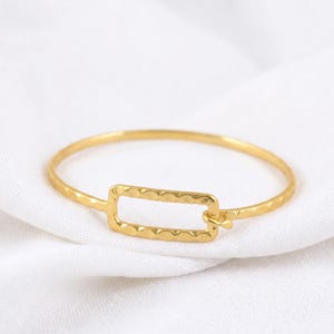 May include: A gold-colored bangle bracelet with a rectangular hammered design in the center. The bracelet is on a white surface. The bracelet is a simple, elegant piece of jewelry.