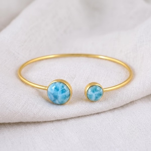 Könnte beinhalten: Ein goldfarbenes, offenes Armband mit zwei runden, polierten Larimar-Edelsteinen. Der größere Stein befindet sich an einem Ende und ein kleinerer Stein am anderen, beide mit einem marmorierten himmelblauen und weißen Muster in einer glatten Fassung.