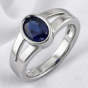 May include: A silver-toned ring featuring a central oval-cut deep blue gemstone set in a smooth bezel. The band has a split-shank design that tapers toward the back, creating a polished and reflective metallic surface around the dark blue stone.