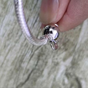 Puede incluir: Una pulsera plateada con un dise&ntilde;o de cadena de serpiente. La pulsera est&aacute; sujeta por una persona. El cierre es una cuenta redonda y brillante con un peque&ntilde;o dije colgante. La pulsera es de metal brillante.