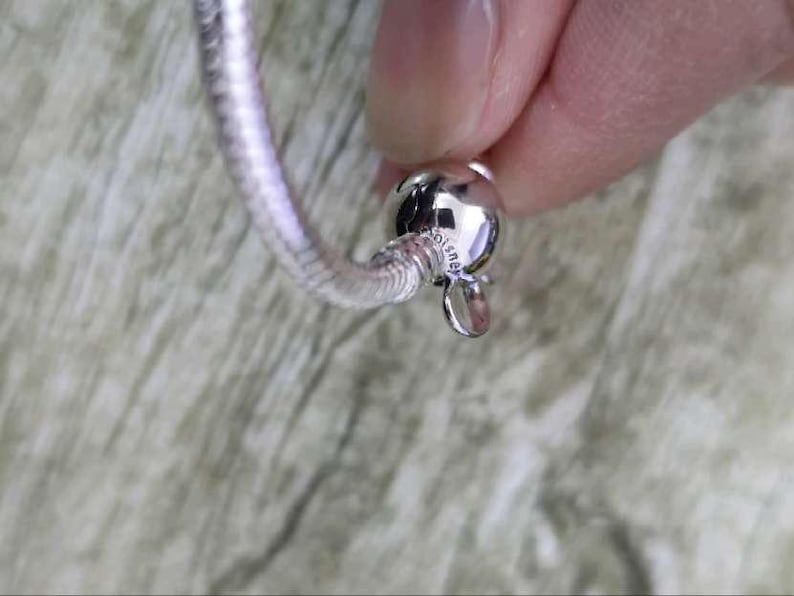 Puede incluir: Una pulsera plateada con un dise&ntilde;o de cadena de serpiente. La pulsera est&aacute; sujeta por una persona. El cierre es una cuenta redonda y brillante con un peque&ntilde;o dije colgante. La pulsera es de metal brillante.