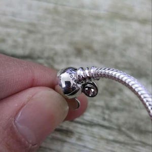 Puede incluir: Primer plano de una pulsera plateada con un cierre redondo y un dise&ntilde;o de cadena de serpiente. El cierre presenta un peque&ntilde;o dije redondo con la palabra "Pandora" grabada. La pulsera es de un material met&aacute;lico brillante.