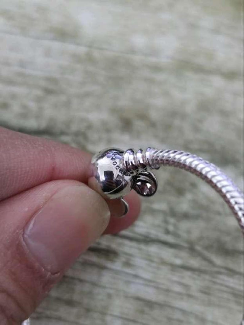 Puede incluir: Primer plano de una pulsera plateada con un cierre redondo y un dise&ntilde;o de cadena de serpiente. El cierre presenta un peque&ntilde;o dije redondo con la palabra "Pandora" grabada. La pulsera es de un material met&aacute;lico brillante.