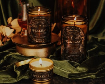 May include: Three candles in glass jars with gold lids. The candles are labelled "Phantom Row" and "Speakeasy". The candles are on a green velvet surface.