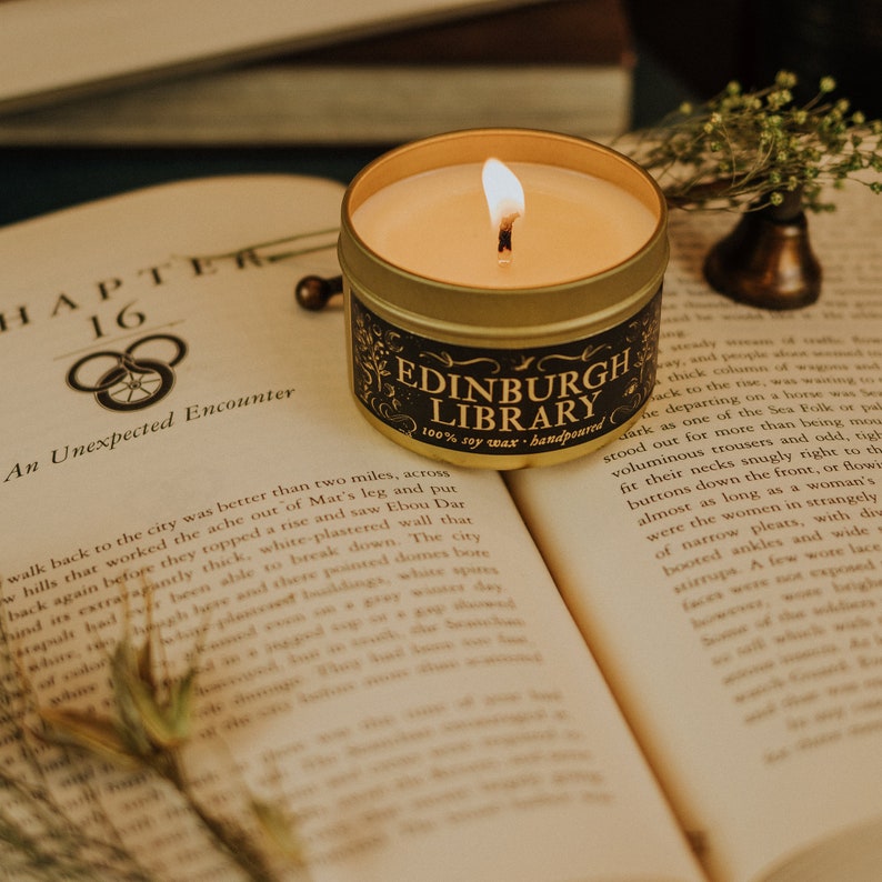 Lit Edinburgh Library 3.3 oz gold tin soy candle sitting on top of open book arranged with a bronze candle snuffer and dried florals