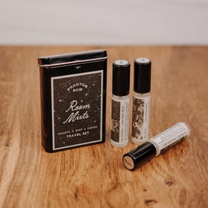 Puede incluir: Un set de viaje Phantom Row Room Mists negro, con una lata rectangular y tres botellas roll-on. La lata dice "Room Mists" y "Balance + Root + Soothe". Las botellas roll-on tienen tapas negras y etiquetas blancas.