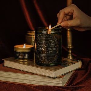 Lifestyle shot of lit Edinburgh Library 11 oz soy candle with a vintage green glass jar, cork top, and 3.3 oz gold tin candle arranged on a stack of book with a brass candlestick holder in the background