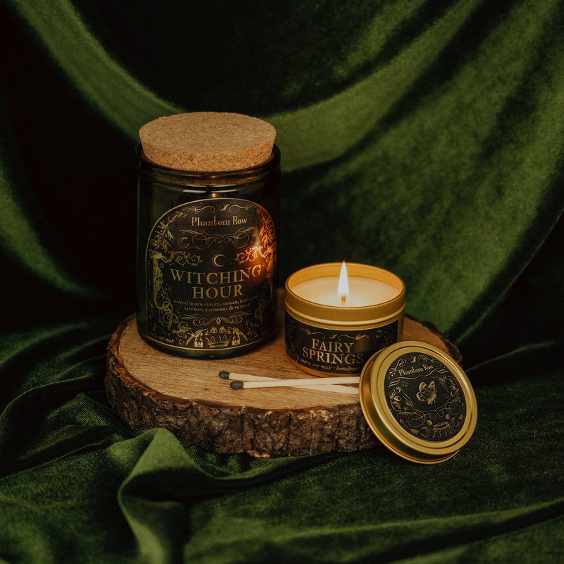 Lifestyle shot of lit Witching Hour 11 oz soy candle with a vintage green glass jar, cork top, and 3.3 oz gold tin candle arranged on a wood stand with a green velvet background