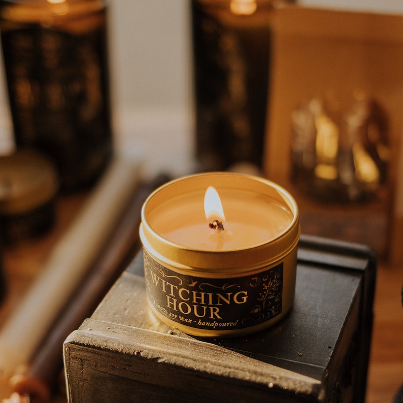Lit Witching Hour 3.3 oz gold candle tin sitting on a black box bathed in golden hour light