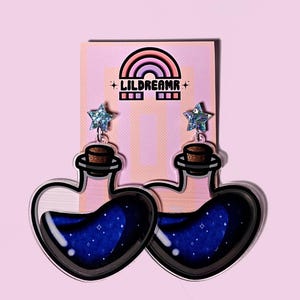 May include: A pair of heart-shaped blue potion bottle earrings with a silver glitter star charm on top. The earrings are made of acrylic and have a black outline.