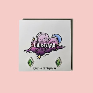 May include: A pair of green gemstone earrings with a silver setting. The earrings are displayed on a white card with a purple and pink cloud illustration and the text "Lil Dreamr" and "Keep on Dreaming".