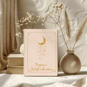 May include: A light peach-colored hardcover journal featuring a gold-toned graphic of a crescent moon with hanging stars and the text "Dripping in Starlight & Moonbeams" printed in a cursive font, enclosed within a decorative rectangular border.