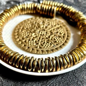 May include: A gold-toned bracelet made of multiple small, linked, textured, round pieces. The bracelet is displayed on a white plate with a decorative, gold-toned, circular design.
