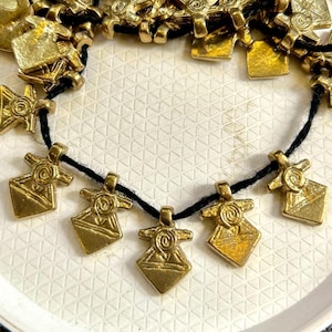 以下が含まれることがあります： 黒い紐と、円形のデザインが施されたピラミッド型の金色のペンダントチャームが複数付いた、金色のネックレス。チャームは白い皿の上に円形に配置されています。