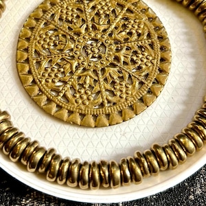 Op de afbeelding: Een gouden ketting met een grote, ronde, sierlijke hanger. De hanger heeft een delicaat, ingewikkeld ontwerp en is gemaakt van messing. De ketting is gemaakt van kleine, ronde kralen.