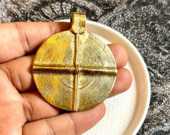 May include: A round, gold-colored pendant with a textured surface and a loop at the top. The pendant has a cross-shaped design in the center.