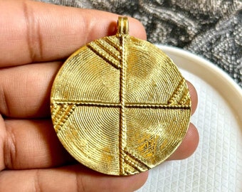 Op de afbeelding: Een goudkleurige, cirkelvormige hanger met een getextureerd oppervlak. De hanger heeft een kruisvormig ontwerp met ingewikkelde patronen en een kleine lus aan de bovenkant voor een ketting. Het ontwerp is verdeeld in kwadranten met gedetailleerde lijnen.