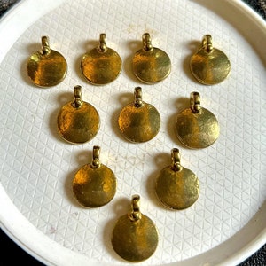 Może przedstawiać: Dziesięć złotych okrągłych zawieszek z pętelką u góry. Zawieszki są ułożone w kółko na białym talerzu.