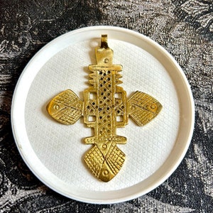 Peut inclure: Un pendentif en métal doré en forme de croix avec des motifs géométriques complexes. La croix est sur un fond blanc.