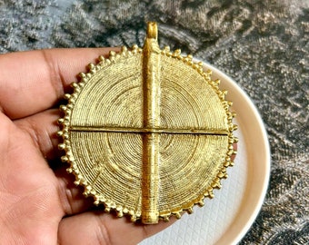 May include: A gold-colored pendant with a circular design featuring concentric circles and a cross-shaped overlay. The pendant has a textured surface and a loop at the top for hanging. The pendant is held in a hand, resting on a white plate.