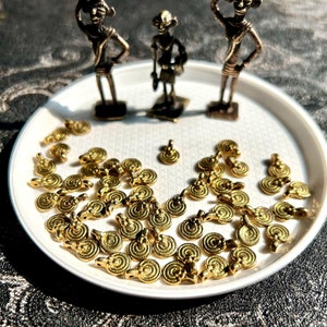 Puede incluir: Un plato blanco con una colección de abalorios de metal dorado. Los abalorios son redondos y tienen un diseño en espiral. Hay tres pequeñas figuritas en el plato, dos están de pie y una está sentada.