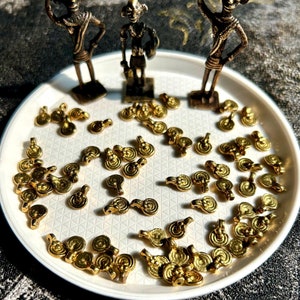 May include: A white plate with a collection of gold-colored metal charms. The charms are shaped like a circle with a small loop on the top. There are three small bronze figurines on the plate, each holding a small object.