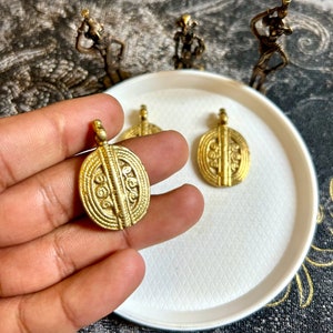 Pode incluir: Três pingentes de metal dourado com designs intrincados. Os pingentes são de forma oval e têm um padrão texturizado em relevo.