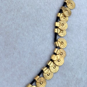 May include: A gold necklace with a black cord and round, coin-shaped charms. The charms have a textured, concentric circle design.