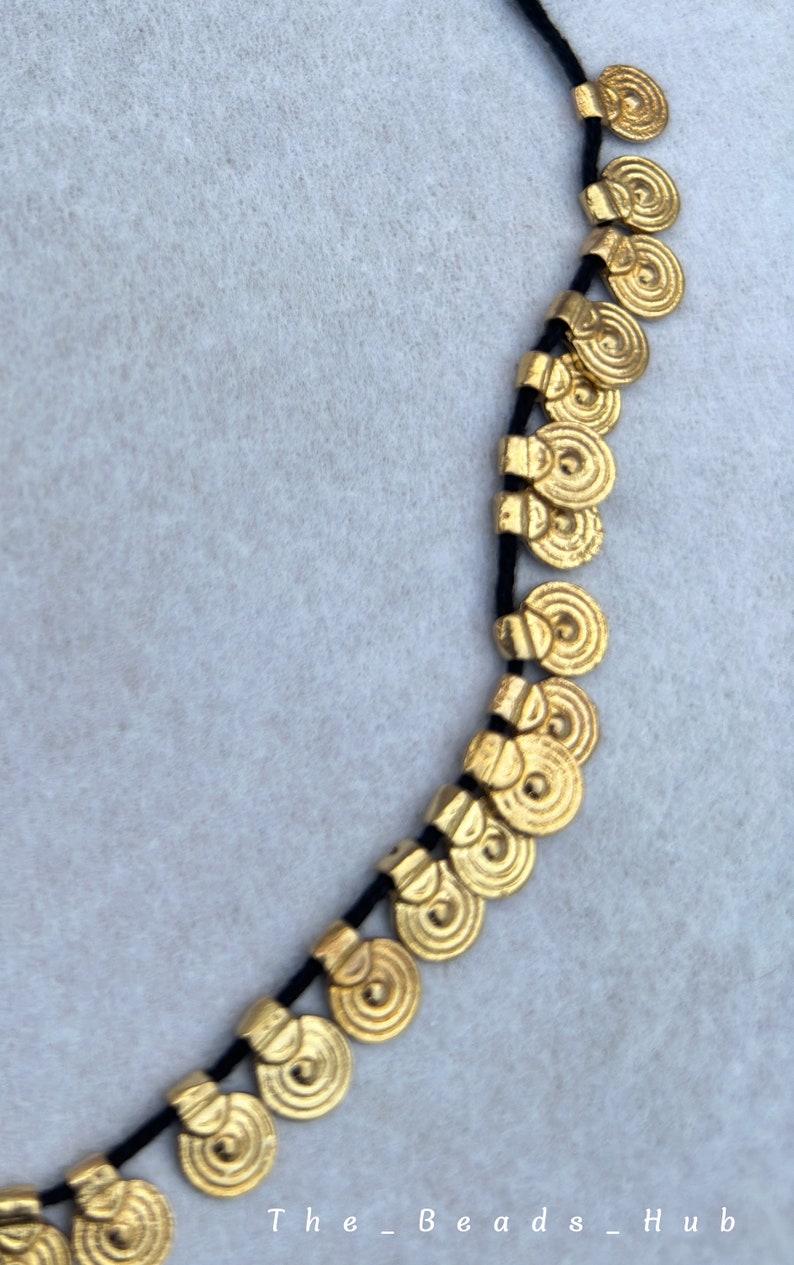 May include: A gold necklace with a black cord and round, coin-shaped charms. The charms have a textured, concentric circle design.