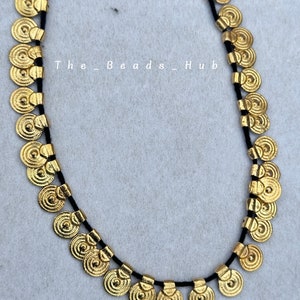 May include: A gold necklace with a black cord and round, coin-shaped charms. The charms have a textured, concentric circle design.