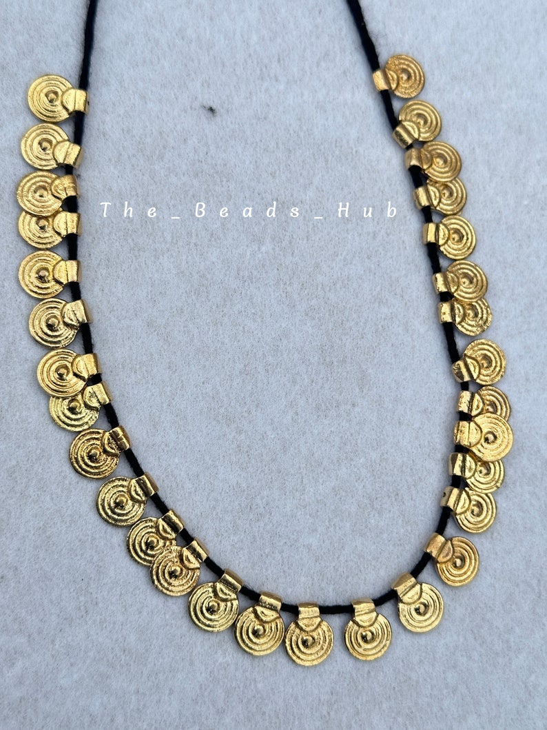 May include: A gold necklace with a black cord and round, coin-shaped charms. The charms have a textured, concentric circle design.
