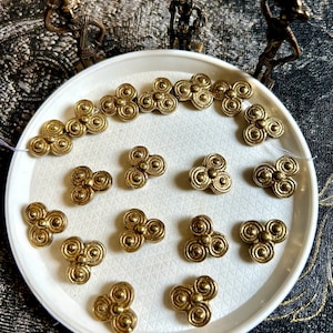 Può includere: Una collezione di 18 perline in metallo dorato con un design a trifoglio. Ogni perlina ha tre lobi arrotondati con un motivo a cerchi concentrici.