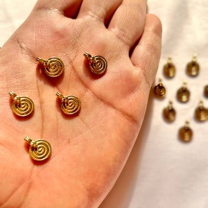 Può includere: Una mano che tiene un gruppo di ciondoli in metallo dorato con un design a spirale. Ci sono altri ciondoli sullo sfondo.