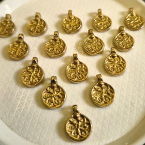 以下が含まれることがあります： 小さな金色のペンダントのコレクション。装飾的で渦巻くデザインが施されています。各ペンダントの上部には、紐を通すためのループが付いています。ペンダントは、白くテクスチャ加工された表面に配置されています。