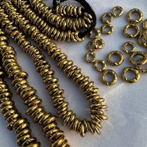 Könnte beinhalten: Nahaufnahme einer goldfarbenen Halskette aus kleinen, runden Perlen. Die Perlen sind spiralförmig angeordnet und auf einer schwarzen Schnur aufgefädelt. Es liegen auch lose Perlen um die Halskette herum. Der Text "The_Beads_Hub" ist in der rechten unteren Ecke des Bildes sichtbar.