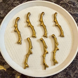 May include: Eight gold-colored, twig-shaped decorative elements arranged on a white, textured plate. The elements have a detailed, organic design, suggesting a natural or botanical theme. The plate is round with a slightly raised edge.