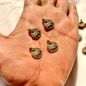 May include: Four small, gold-colored, round pendants with a textured design are displayed in a hand. Each pendant has a loop at the top for stringing. The pendants have a detailed pattern of dots and curved lines.