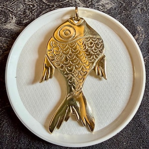 May include: A gold-coloured fish pendant with detailed scale and fin designs. The pendant is suspended from a small loop and rests on a white, textured plate. The background is a dark, patterned fabric.