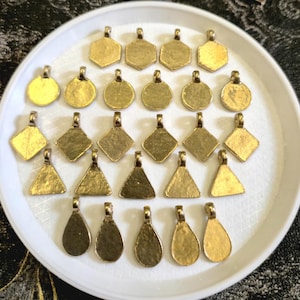 May include: A selection of brass pendants in diverse forms: hexagons, circles, diamonds, triangles, and teardrops. Each pendant features a loop for attaching to a chain. The pendants are displayed on a white plate.